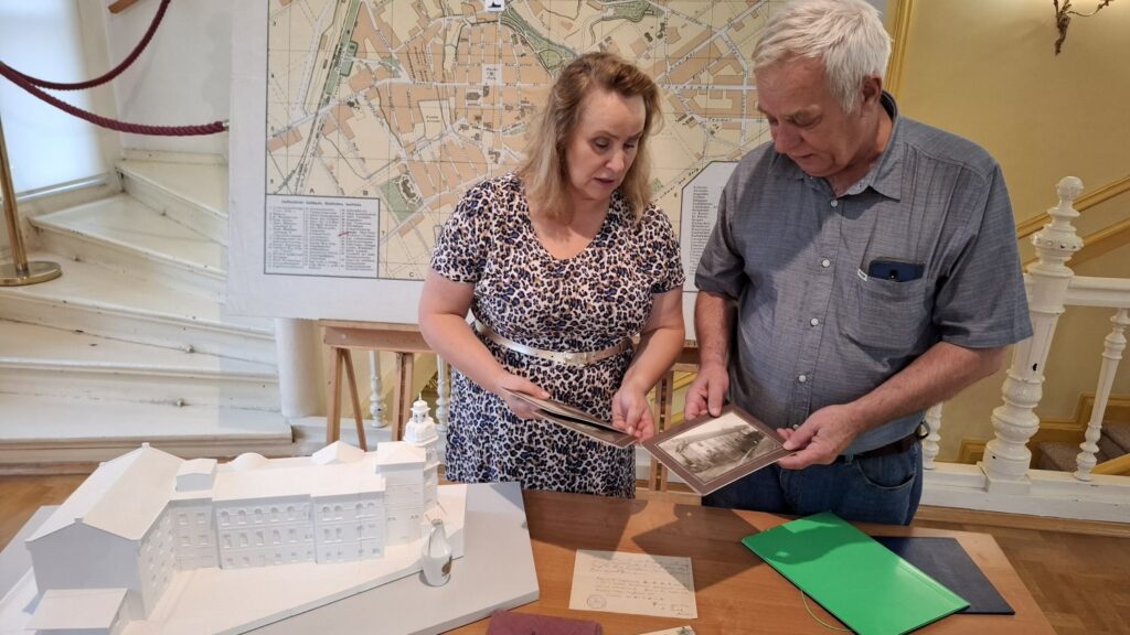 Dwie osoby stoją za stołem, na którym znajdują się archiwalne dokumenty i przeglądają stare fotografie przekazane do Muzeum. W tle klatka schodowa i wielkoformatowa mapa Koszalina (kliknięcie spowoduje powiększenie zdjęcia).