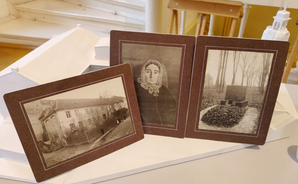 Na stole, przed makietą Muzeum w Koszalinie stoją trzy, czarno-białe, archiwalne fotografie. Widać na nich: zabudowania, starszą kobietę i nagrobek (kliknięcie spowoduje powiększenie zdjęcia).