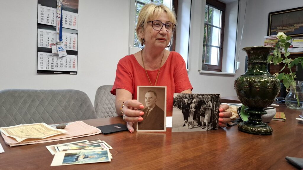 Kobieta siedzi przy stole, w biurze, i pokazuje do obiektywu dwie archiwalne, rodzinne fotografie (kliknięcie spowoduje powiększenie zdjęcia).