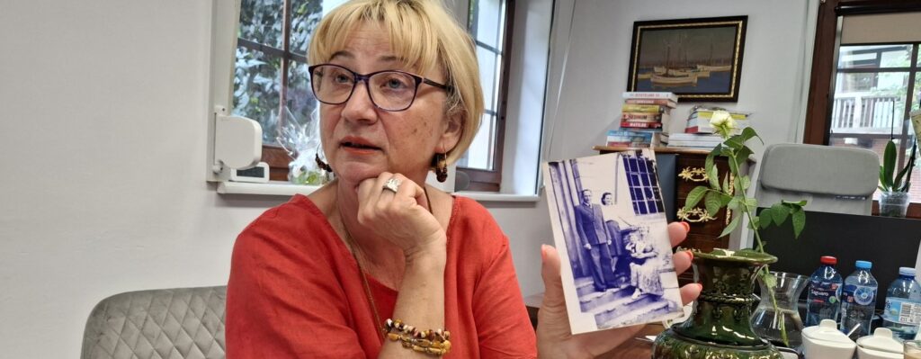 Kobieta siedzi przy stole, w biurze, i trzyma w dłoni archiwalne zdjęcie rodzinne, które pokazuje do obiektywu (kliknięcie spowoduje powiększenie zdjęcia).