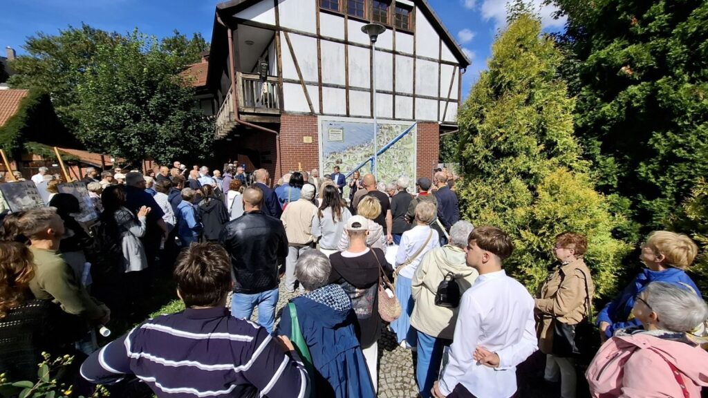 Ludzie stoją na dziedzińcu muzeum i uczestniczą w otwarciu wystawy. W tle budynek w formie szachulcowej, na którego ścianie wisi mapa "Wędrownik koszaliński" (kliknięcie spowoduje powiększenie zdjęcia).