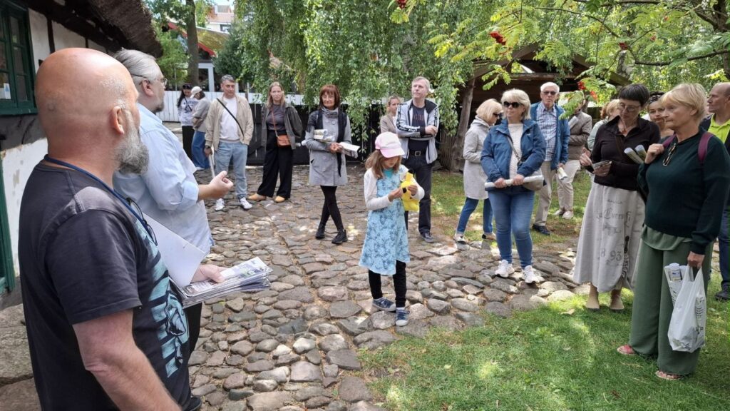 Ludzie stoją przed zabytkową chałupą, przed wejściem i na trawniku. Uczestniczą w oprowadzaniu po wystawie (kliknięcie spowoduje powiększenie zdjęcia).