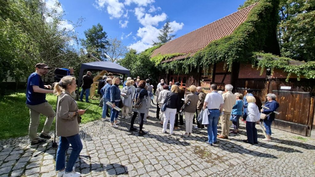 Ludzie stoją przed budynkiem stodoły i uczestniczą w oprowadzaniu po wystawie. Po lewej i po prawej widoczne zabudowania. W tle namiot, pod którym znajduje się model szkieletu szachulcowego (kliknięcie spowoduje powiększenie zdjęcia).