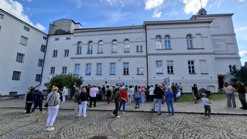 Ludzie stoją przed budynkiem muzeum i chodzą po dziedzińcu. W tle widoczny budynek muzeum (kliknięcie spowoduje powiększenie zdjęcia).