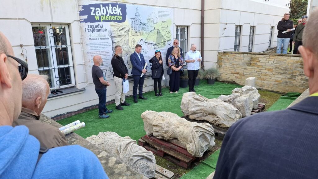 Ludzie stoją przed ścianą budynku muzeum, przy której leżą fragmenty pomnika Fryderyka Wilhelma I. Jedna osoba mówi do mikrofonu (kliknięcie spowoduje powiększenie zdjęcia).