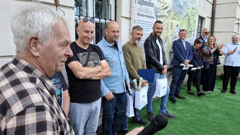 Grupka ludzi stoi przed ścianą muzeum i się uśmiecha. Za ich plecami wisi baner wystawy "Zabytek w plenerze" (kliknięcie spowoduje powiększenie zdjęcia).