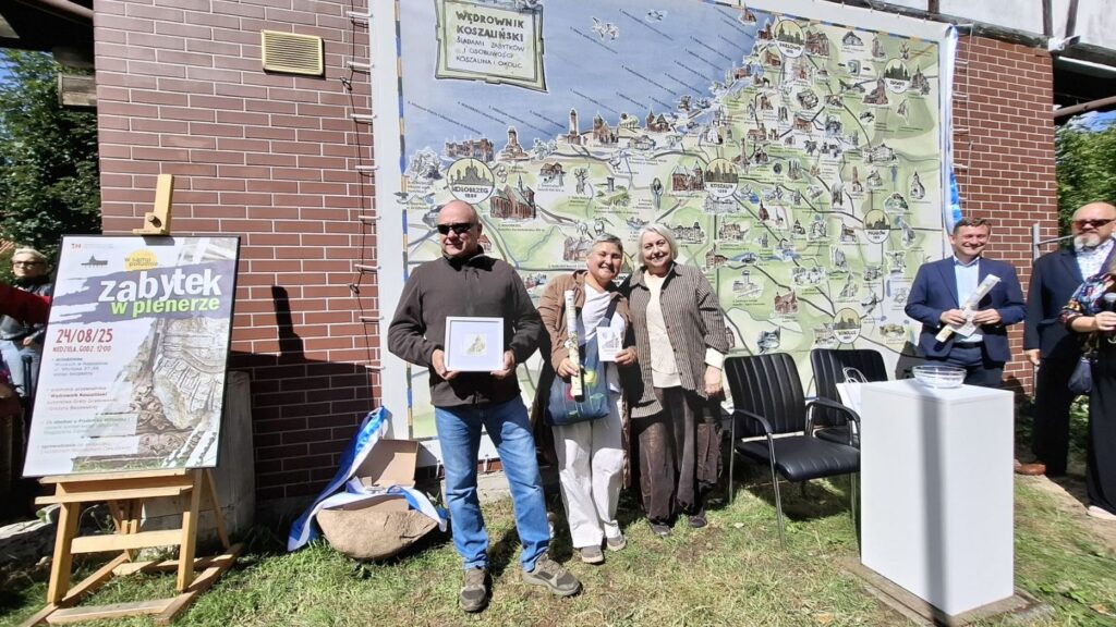 Pięć osób stoi przed ścianą budynku, na tle mapy "Wędrownik koszaliński" i pozuje do zdjęcia. Po prawej stronie stoi oprawiony w ramę plakat wydarzenia (kliknięcie spowoduje powiększenie zdjęcia).