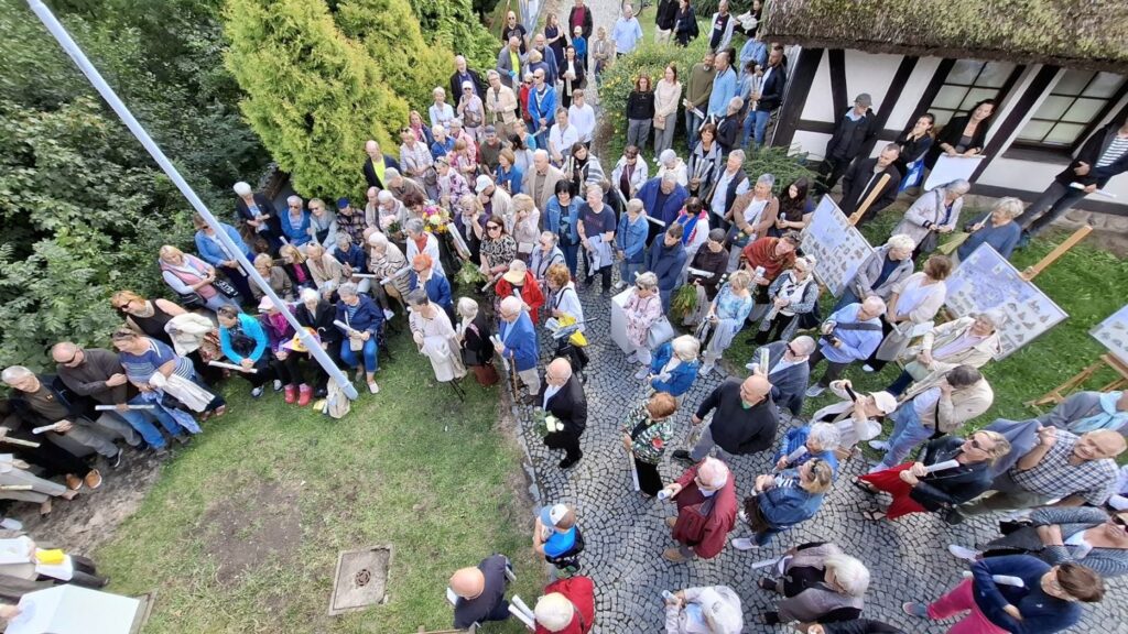 Ludzie stoją pod budynkiem krytym strzechą i słuchają przemówienia osób otwierających wydarzenie (kliknięcie spowoduje powiększenie zdjęcia).