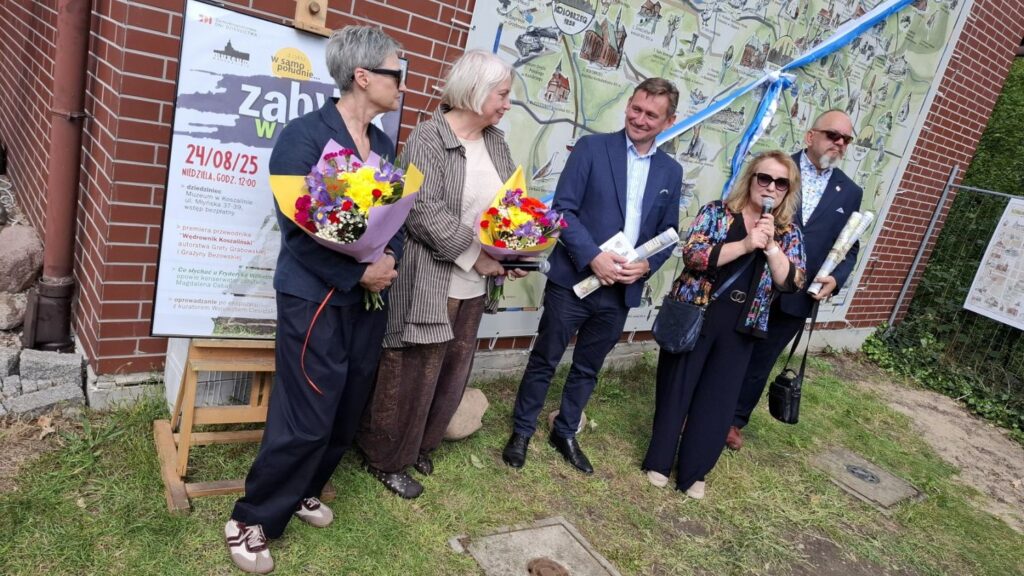 Pięć osób stoi przed ścianą budynku, na tle mapy "Wędrownik koszaliński". Jedna z osób mówi do mikrofonu, dwie kobiety trzymają w rękach bukiet kwiatów (kliknięcie spowoduje powiększenie zdjęcia).