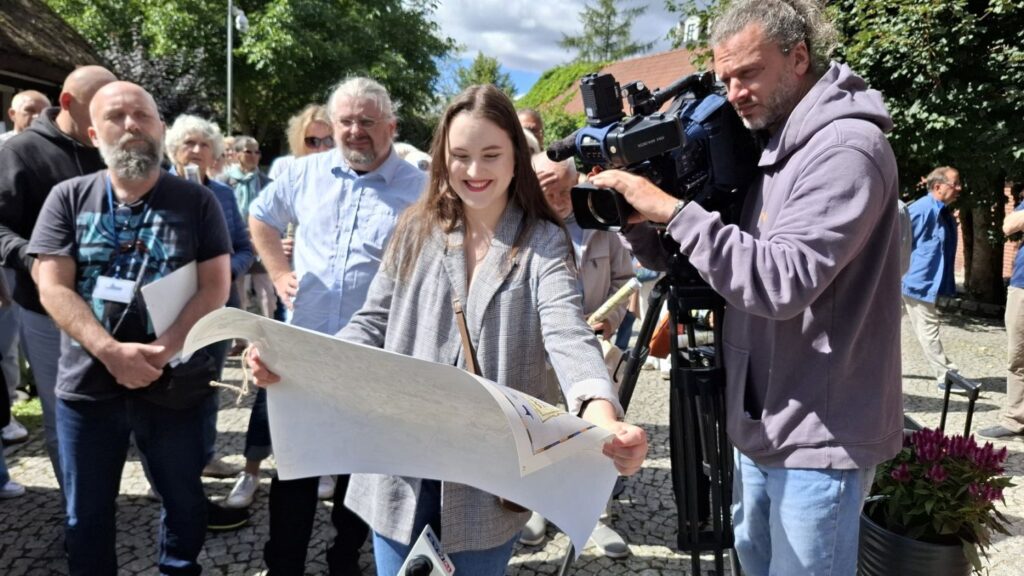 Kobieta trzyma w rękach rozwiniętą mapę. Za nią stoi operator kamery i robi ujęcie. W tle ludzie zgromadzeni na terenie muzeum (kliknięcie spowoduje powiększenie zdjęcia).