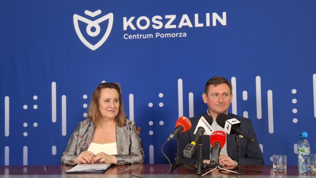 Dwie osoby siedzą przy stole, przed mikrofonami. W tle niebieska plansza z napisem Koszalin Centrum Pomorza (kliknięcie spowoduje powiększenie zdjęcia).
