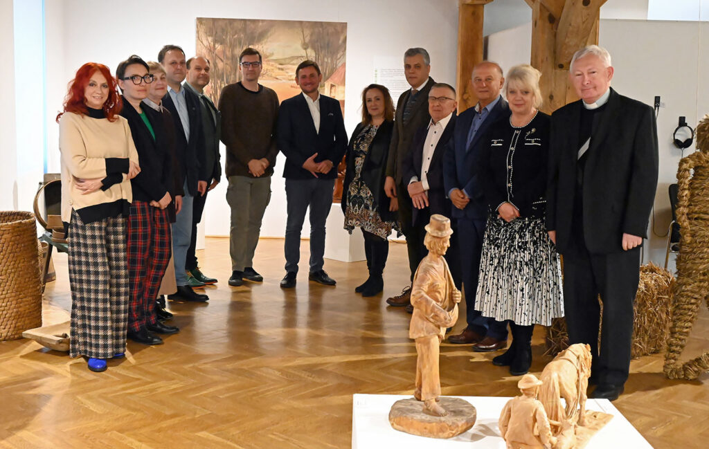 Grupa osób pozuje do wspólnego zdjęcia; w tle wystawa muzealna. Po prawej stronie i z przodu widoczne zbiory muzealne prezentowane na wystawie.