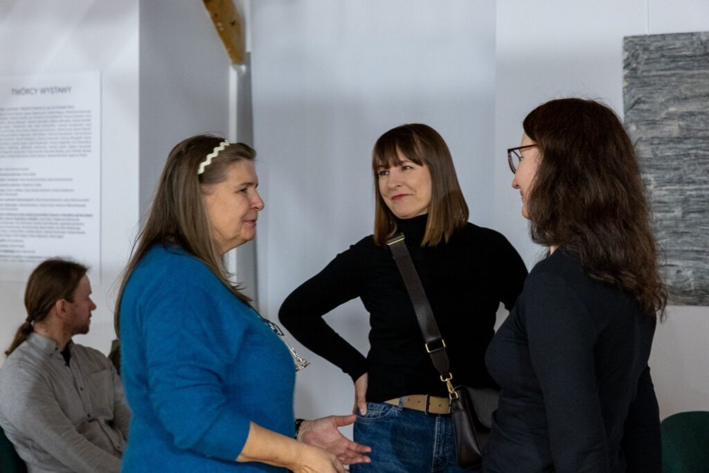 Trzy osoby stoją zwrócone do siebie i ze sobą rozmawiają. W tle jedna osoba siedzi na krześle. Po prawej fragment eksponatu zawieszony na ścianie (kliknięcie spowoduje powiększenie zdjęcia)