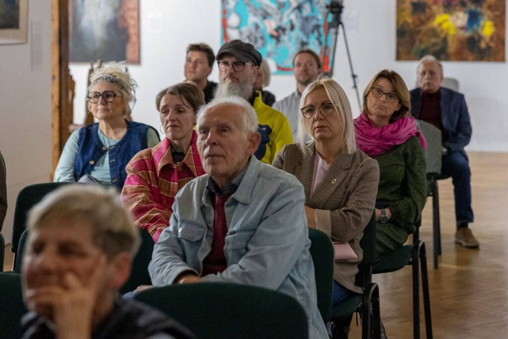 Uczestnicy spotkania siedzą na krzesłach i słuchają w skupieniu prezentacji. W tle, na ścianach, wiszą prace malarskie (kliknięcie spowoduje powiększenie zdjęcia)