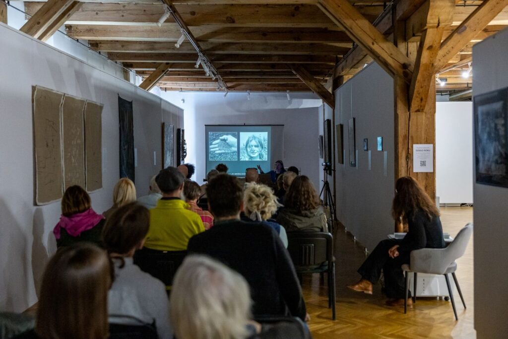 Sala wystawowa z drewnianym stropem, widok na ekran z prezentacją i publiczność oglądającą pokaz. Na ścianach wiszą prace malarskie (kliknięcie spowoduje powiększenie zdjęcia)