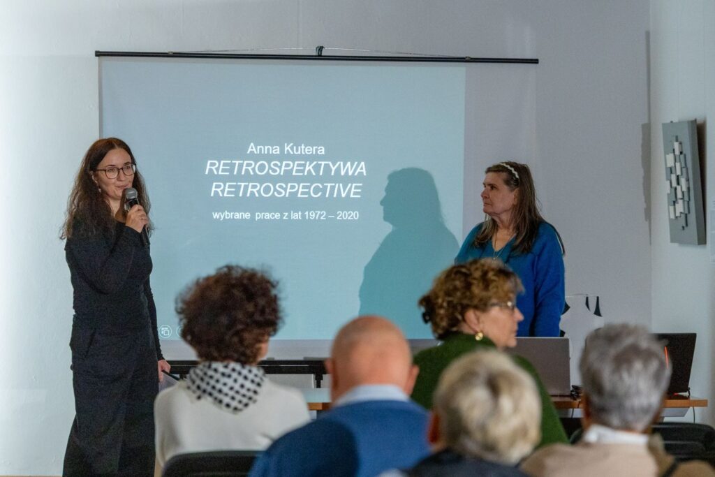 Dwie kobiety stoją przed ekranem z napisem ‘Anna Kutera. Retrospektywa. Wybrane prace z lat 1972–2020’, przemawiają do publiczności (kliknięcie spowoduje powiększenie zdjęcia)