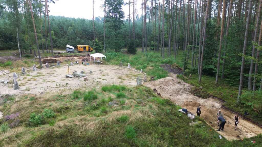 Widok na kamienne kręgi. W tle las i droga leśna, na której stoi samochód. Po lewej stronie cztery osoby pracujące w wykopie (kliknięcie spowoduje powiększenie zdjęcia).