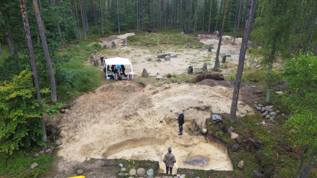 Kamienne kręgi i wykop archeologiczny z lotu ptaka, w którym pracują ludzie. W tle las (kliknięcie spowoduje powiększenie zdjęcia).