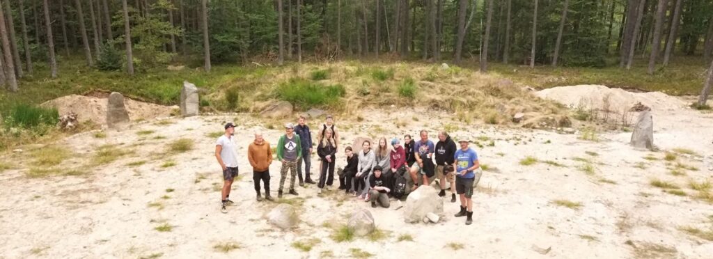 Grupa osób pozuje do wspólnego zdjęcia, na tle kamiennych kręgów i lasu (kliknięcie spowoduje powiększenie zdjęcia).