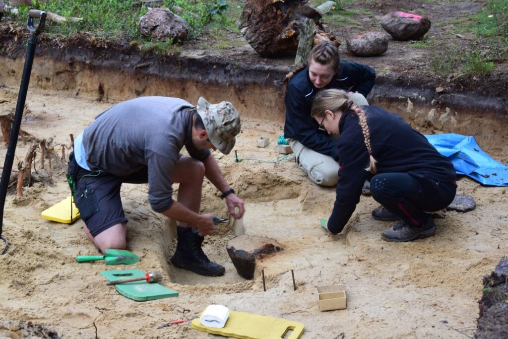 Trzy osoby pracują w wykopie i odsłaniają odnaleziony artefakt (kliknięcie spowoduje powiększenie fotografii).