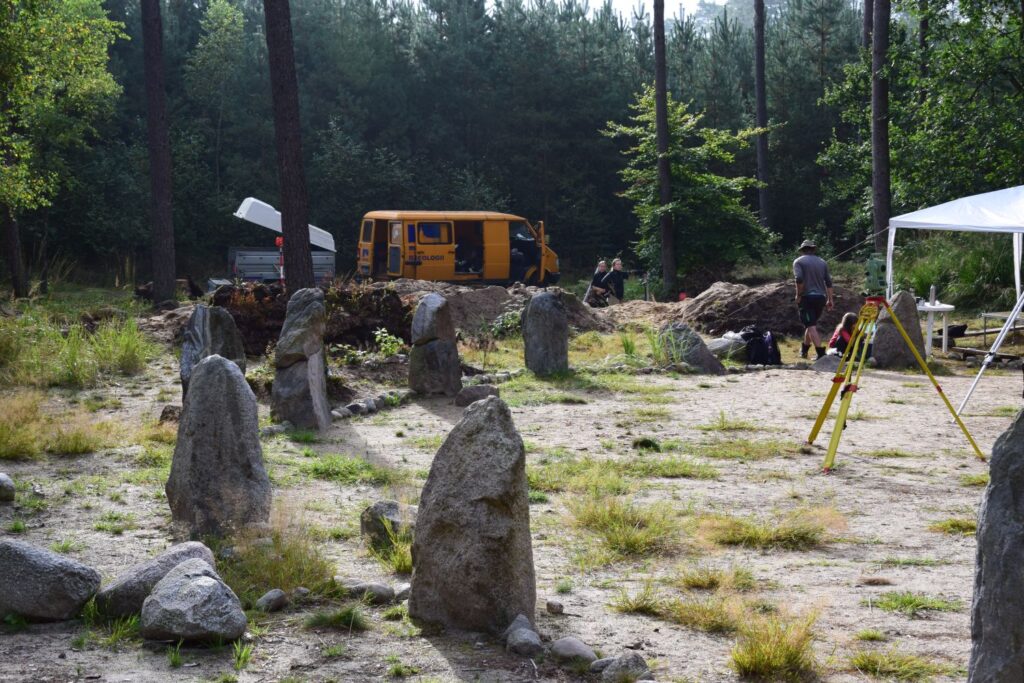 Fragment kamiennych kręgów. W tle las i samochód. Na środku stoi urządzenie pomiarowe, a po prawej stronie idzie jedna osoba (kliknięcie spowoduje powiększenie fotografii).