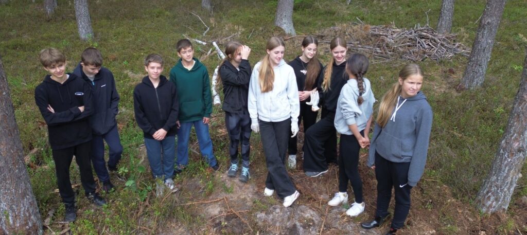 Dziesięcioosobowa grupa ośmioklasistów pozuje do wspólnego zdjęcia. W tle pnie drzew i ściółka leśna (kliknięcie spowoduje powiększenie zdjęcia).