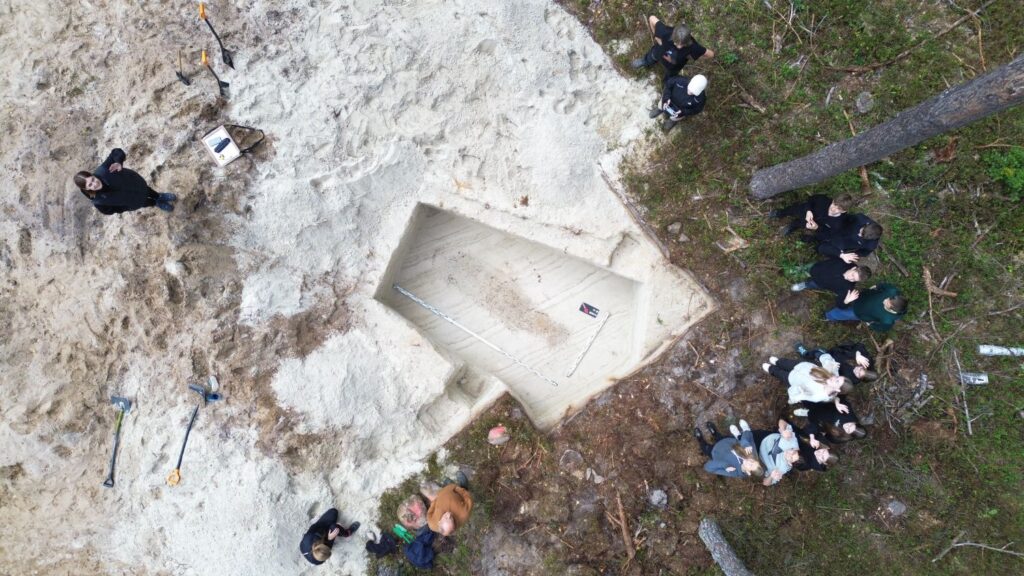 Wykop archeologiczny z drona. Wokół stoi grupa ośmioklasistów, która się uśmiecha i macha do obiektywu (kliknięcie spowoduje powiększenie zdjęcia).