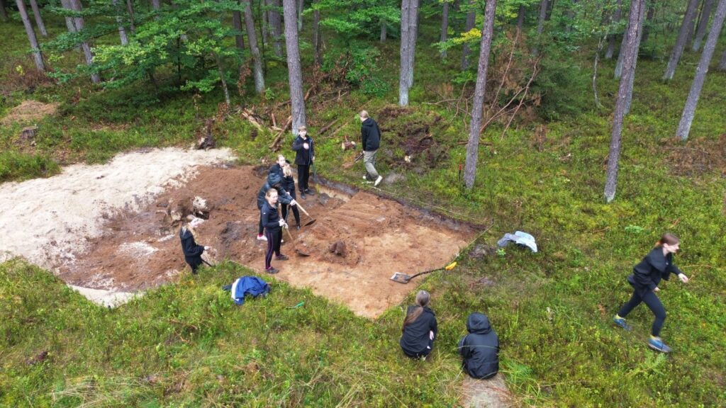 Grupa ośmioklasistów pracuje w wykopie archeologicznym. W tle las (kliknięcie spowoduje powiększenie zdjęcia).