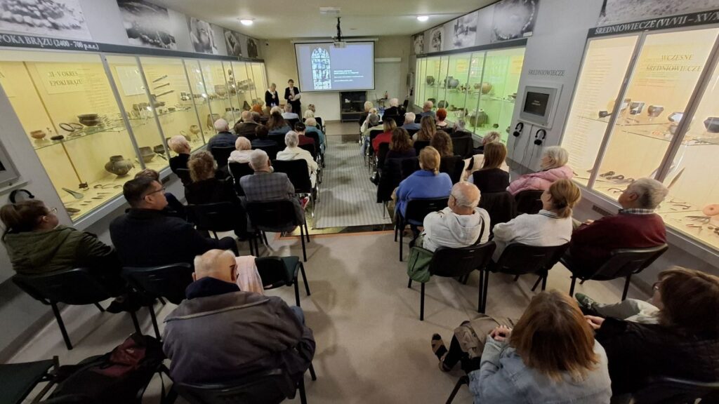 Uczestnicy spotkania siedzący w sali muzealnej i słuchający wykładu o witrażach, w tle gabloty z eksponatami archeologicznymi (kliknięcie powiększy zdjęcie).