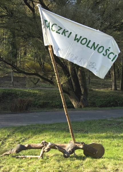 Fotografia przedstawiająca drewnianą konstrukcję z białą flagą z napisem „TACZKI WOLNOŚCI” ustawioną na trawie w parku (kliknięcie powiększy zdjęcie).