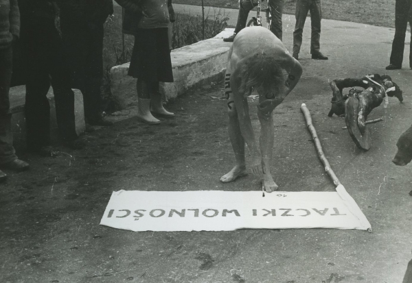 Czarno-biała fotografia przedstawiająca nagiego mężczyznę pochylającego się nad materiałem z napisem „TACZKI WOLNOŚCI” rozłożonym na ziemi. W tle stoją obserwujące osoby oraz elementy instalacji (kliknięcie powiększy zdjęcie).