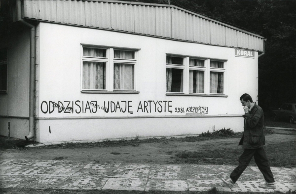 Czarno-biała fotografia przedstawiająca mężczyznę stojącego pośród dużych kamieni i unoszącego ręce nad głową. W tle na trawie stoi druga osoba, a za nimi widoczny jest pas drzew (kliknięcie powiększy zdjęcie).