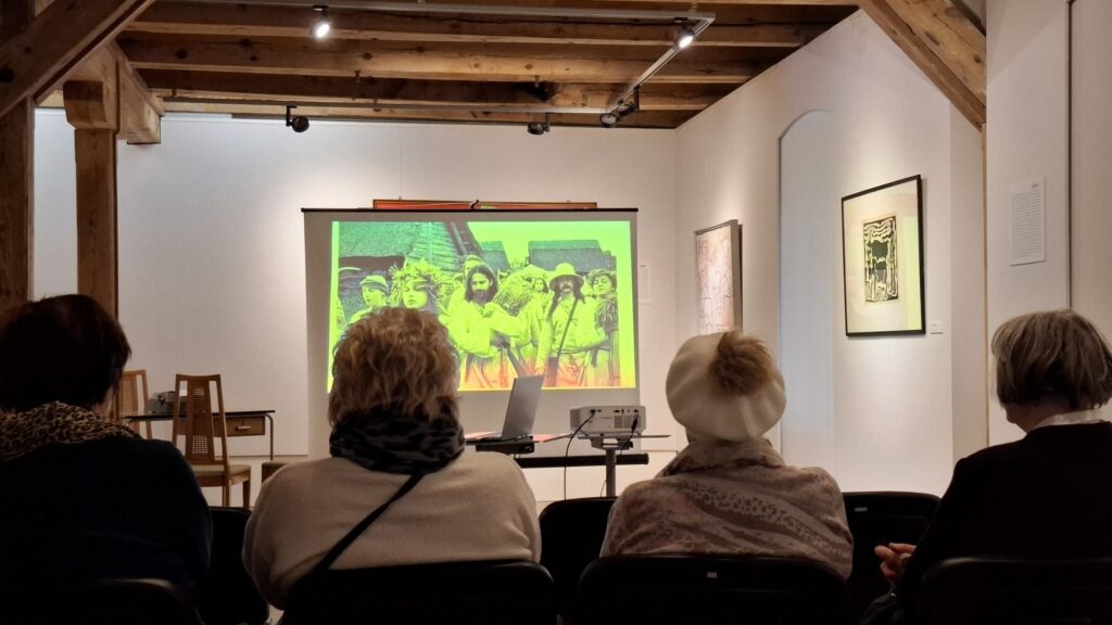 Ludzie siedzą na sali wystawowej i słuchają wykładu. W tle wyświetla się prezentacja multimedialna. Na ścianach wiszą obrazy - elementy ekspozycji muzealnej (kliknięcie spowoduje powiększenie zdjęcia)