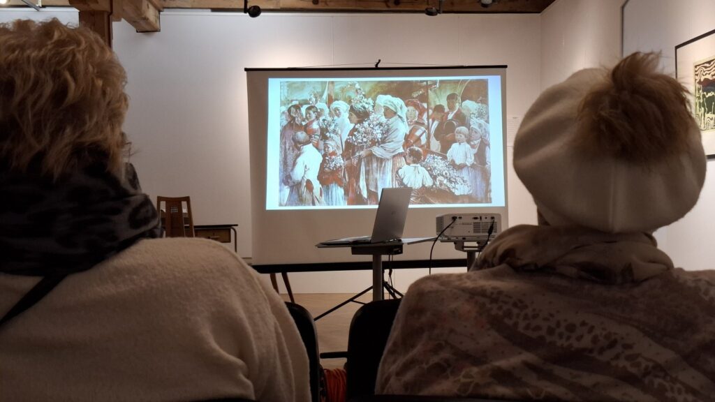 Ludzie siedzą na sali wystawowej i słuchają wykładu. W tle wyświetla się prezentacja multimedialna, a przed nią stoi sprzęt komputerowy (kliknięcie spowoduje powiększenie zdjęcia)