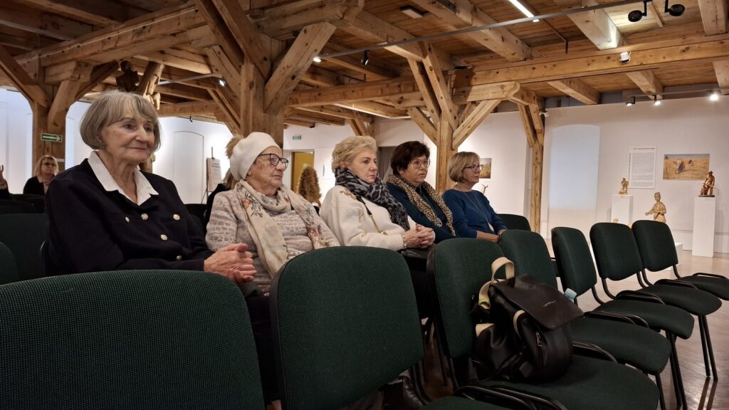 Osoby siedzą na krzesłach i słuchają wykładu. W tle, na ścianach, elementy ekspozycji muzealnej (kliknięcie spowoduje powiększenie zdjęcia)