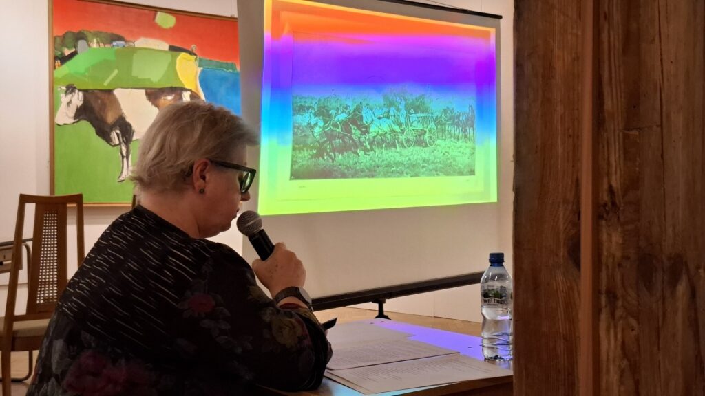 Kobieta siedzi przy biurku i mówi do mikrofonu. W tle wyświetla się prezentacja multimedialna. Po lewej wisi obraz - element ekspozycji na wystawie (kliknięcie spowoduje powiększenie zdjęcia)