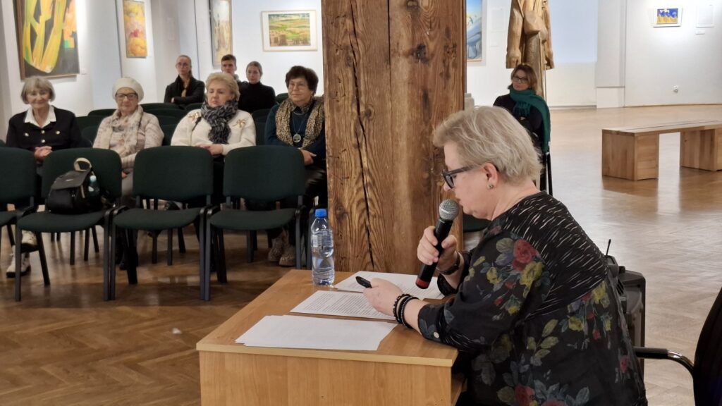 Na pierwszym planie, siedząca przy biurku kobieta, mówi do mikrofonu. Po lewej siedzą ludzie na krzesłach i słuchają wykładu. W tle, na ścianach, elementy ekspozycji muzealnej (kliknięcie spowoduje powiększenie zdjęcia)