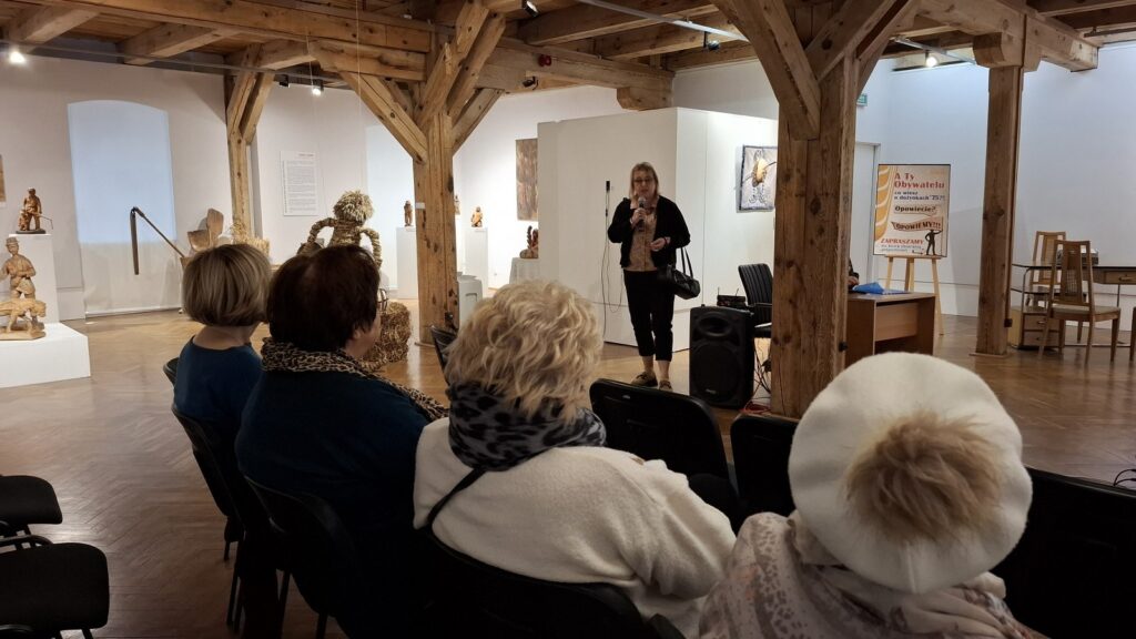 Do ludzi siedzących na krzesłach przemawia jedna osoba. W tle elementy wystawy dożynkowej (kliknięcie spowoduje powiększenie zdjęcia)