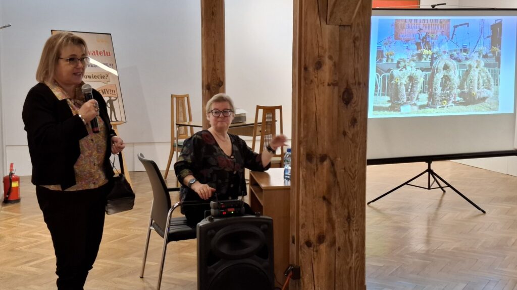 Po lewej stoi kobieta mówiąca do mikrofonu. Obok niej, przed biurkiem, siedzi kobieta i uśmiecha się. Po prawej stoi rozkładana ścianka, na której wyświetlana jest prezentacja (kliknięcie spowoduje powiększenie zdjęcia)