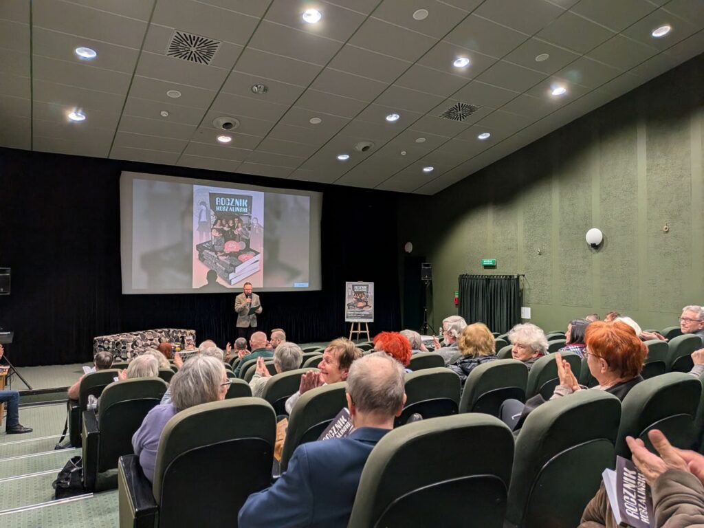 Sala kinowa wypełniona publicznością podczas spotkania autorskiego; na scenie prowadząca osoba stoi przed ekranem z prezentacją (kliknięcie powiększy zdjęcie)