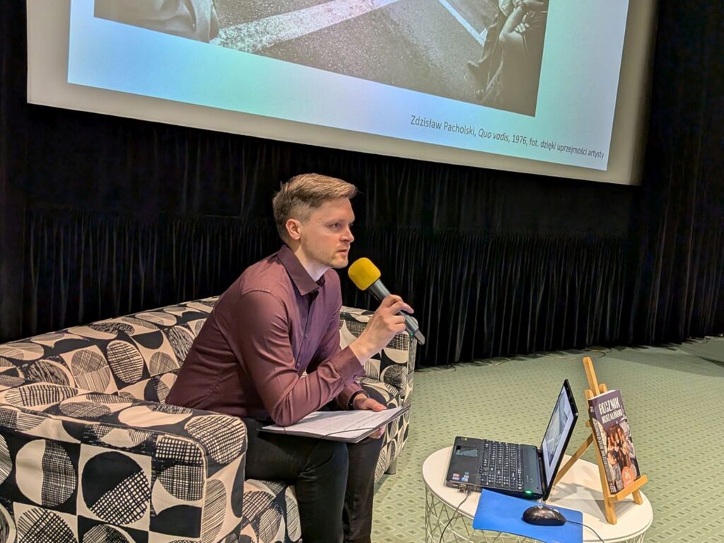 Osoba prowadząca wydarzenie siedzi na fotelu z mikrofonem, obok stolik z laptopem i książką, a na ekranie w tle widoczny slajd prezentacji (kliknięcie powiększy zdjęcie)