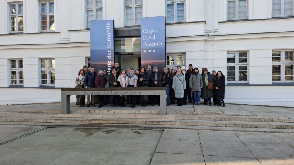 Pracownicy Muzeum w Koszalinie pozująca do zdjęcia przed budynkiem muzeum w Greifswaldzie, na tle elewacji, na której wiszą banery (kliknięcie powiększy zdjęcie).