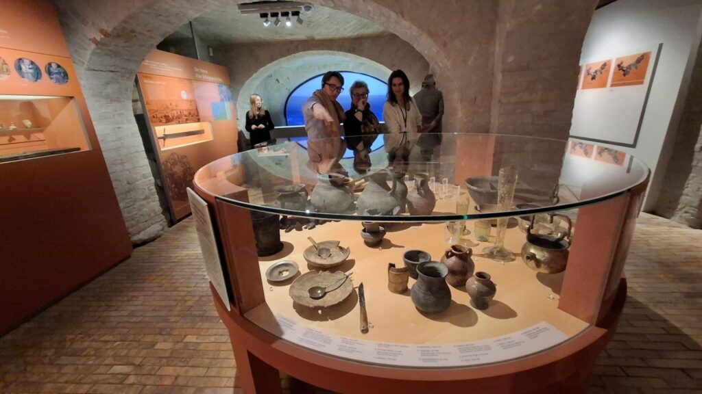 Okrągła gablota z naczyniami ceramicznymi i drobnymi eksponatami, wokół której stoją zwiedzający w muzealnym wnętrzu (kliknięcie powiększy zdjęcie)