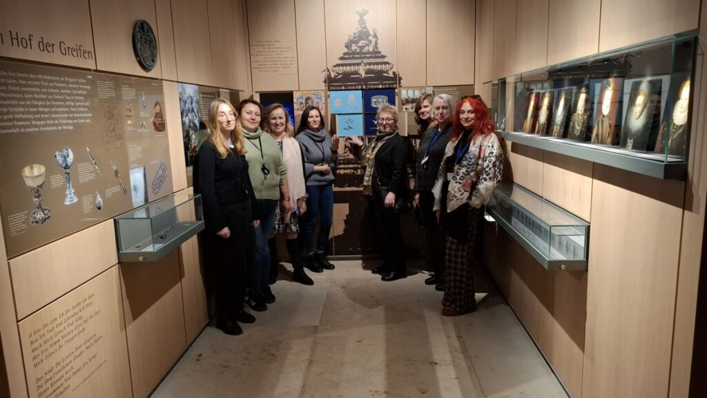 Grupa osób pozuje do zdjęcia w sali wystawowej, pomiędzy gablotami i planszami z opisami ekspozycji (kliknięcie powiększy zdjęcie)