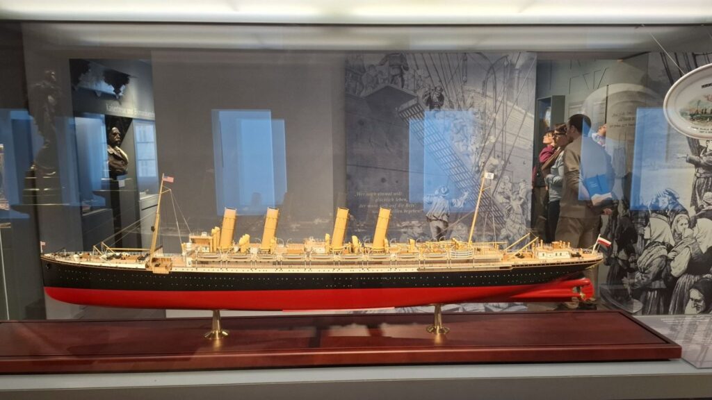 Model statku pasażerskiego w gablocie muzealnej, prezentowany na tle archiwalnych fotografii (kliknięcie powiększy zdjęcie)