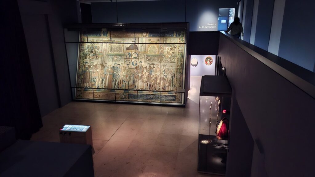 Sala muzealna z dużym historycznym arrasem lub tkaniną ścienną, oglądana z perspektywy wejścia (kliknięcie powiększy zdjęcie)