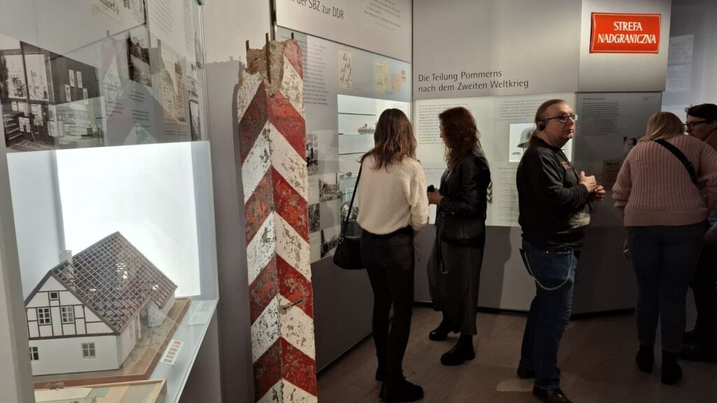 Zwiedzający stoją przy ekspozycji muzealnej z planszami informacyjnymi i obiektami historycznymi (kliknięcie powiększy zdjęcie