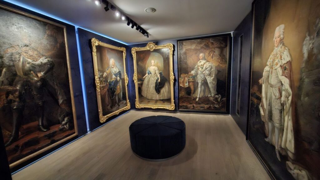 Sala wystawowa z dużymi obrazami olejnymi w złoconych ramach, ustawionymi na ciemnych ścianach (kliknięcie powiększy zdjęcie)