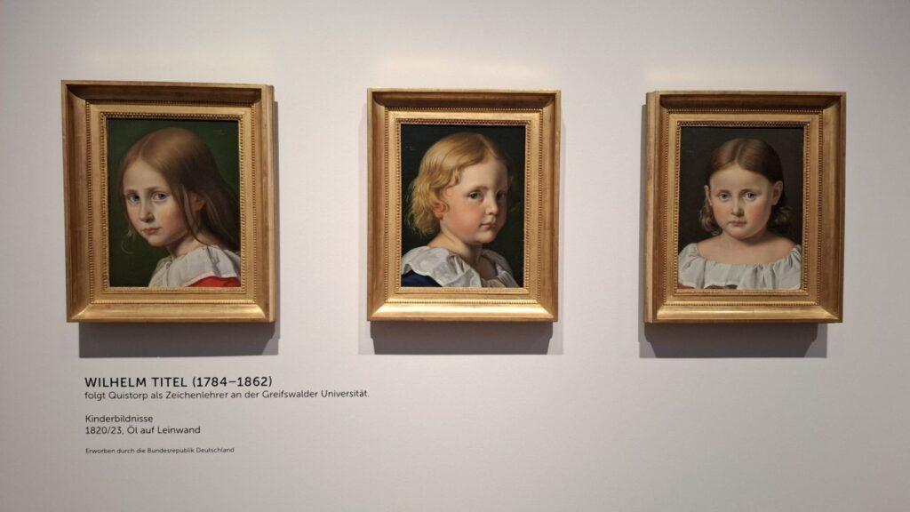 Trzy portrety dzieci w klasycznych ramach, zawieszone obok siebie na ścianie galerii (kliknięcie powiększy zdjęcie)