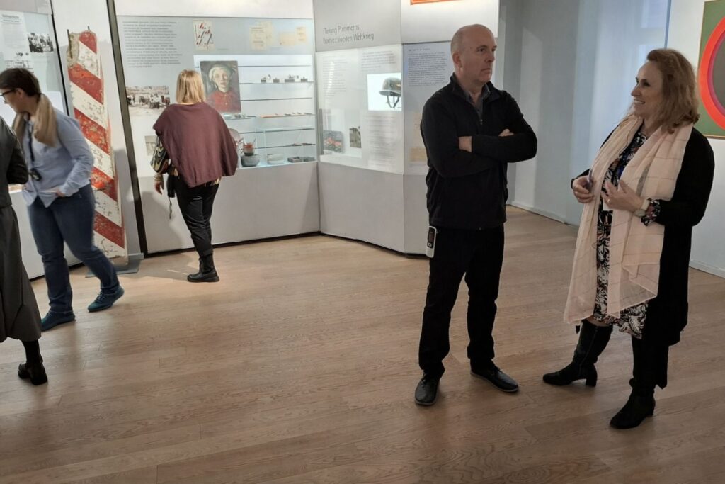 Dwie osoby rozmawiają stojąc w sali muzealnej. Na dalszym planie inni zwiedzający oglądają gabloty i panele informacyjne (kliknięcie powiększy zdjęcie)